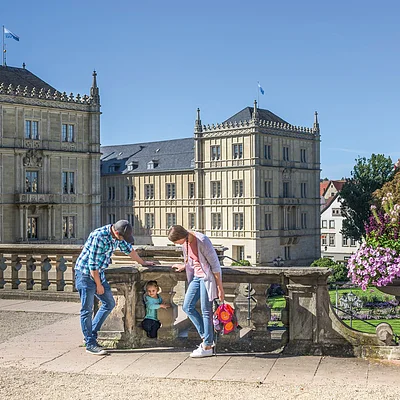 Eine bunt gekleidete Familie ist vor einem Schloss, welches aus mehreren Bauten besteht, zu sehen. Im Hintergrund ist ein Schlossgarten erkennbar.