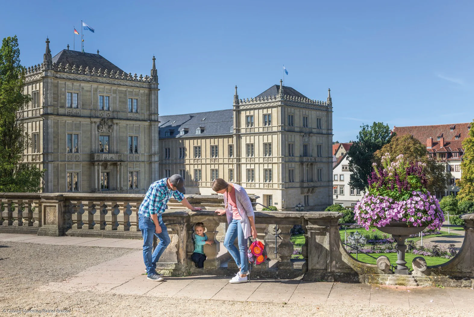 Eine bunt gekleidete Familie ist vor einem Schloss, welches aus mehreren Bauten besteht, zu sehen. Im Hintergrund ist ein Schlossgarten erkennbar.