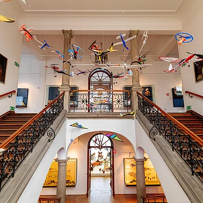 Innenansicht eines Museums mit Treppe, hängenden Modellflugzeugen und Skulpturen von Kindern.