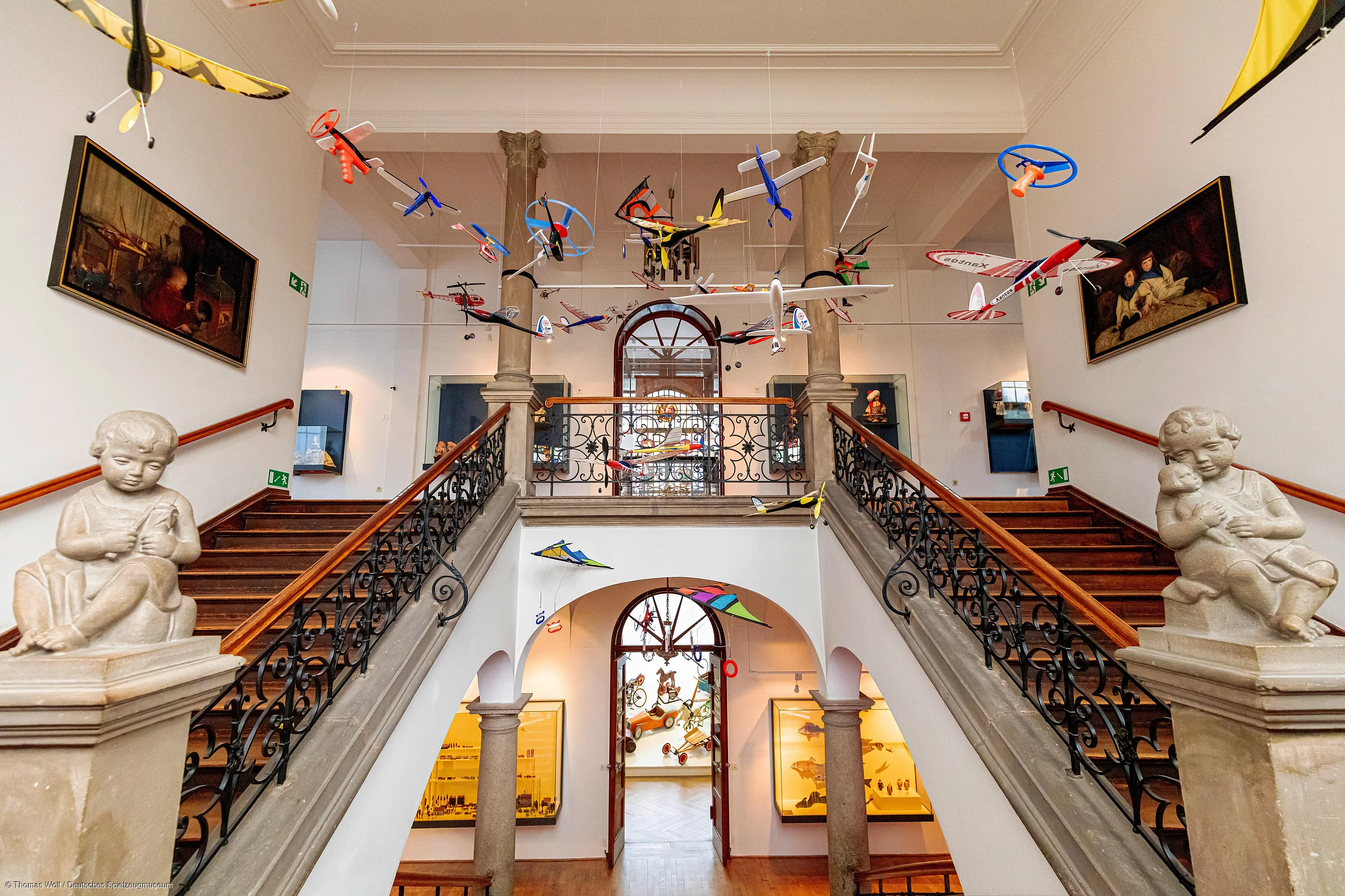 Innenansicht eines Museums mit Treppe, hängenden Modellflugzeugen und Skulpturen von Kindern.