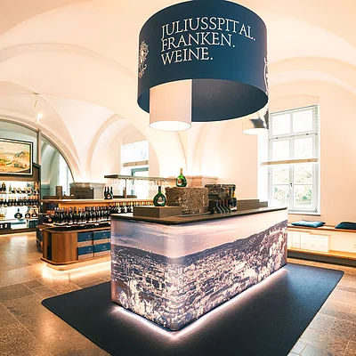 Innenraum eines Verkaufsraums mit Weinflaschen und einem beleuchteten Tresen unter einem runden Schild mit der Aufschrift „Juliusspital Franken Weine“