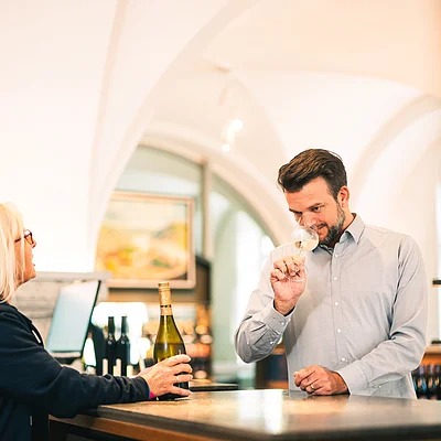Mann riecht an Weinglas, Frau hält Weinflasche an Theke in hellem Raum mit gewölbter Decke