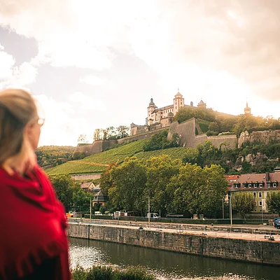 Frau mit rotem Schal blickt auf eine Burg auf einem bewaldeten Hügel über einem Fluss und Häusern.
