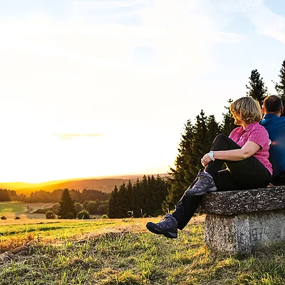 Zwei Personen sitzen auf einer Steinbank und schauen in die untergehende Sonne über einer Landschaft mit Wiesen und Bäumen.