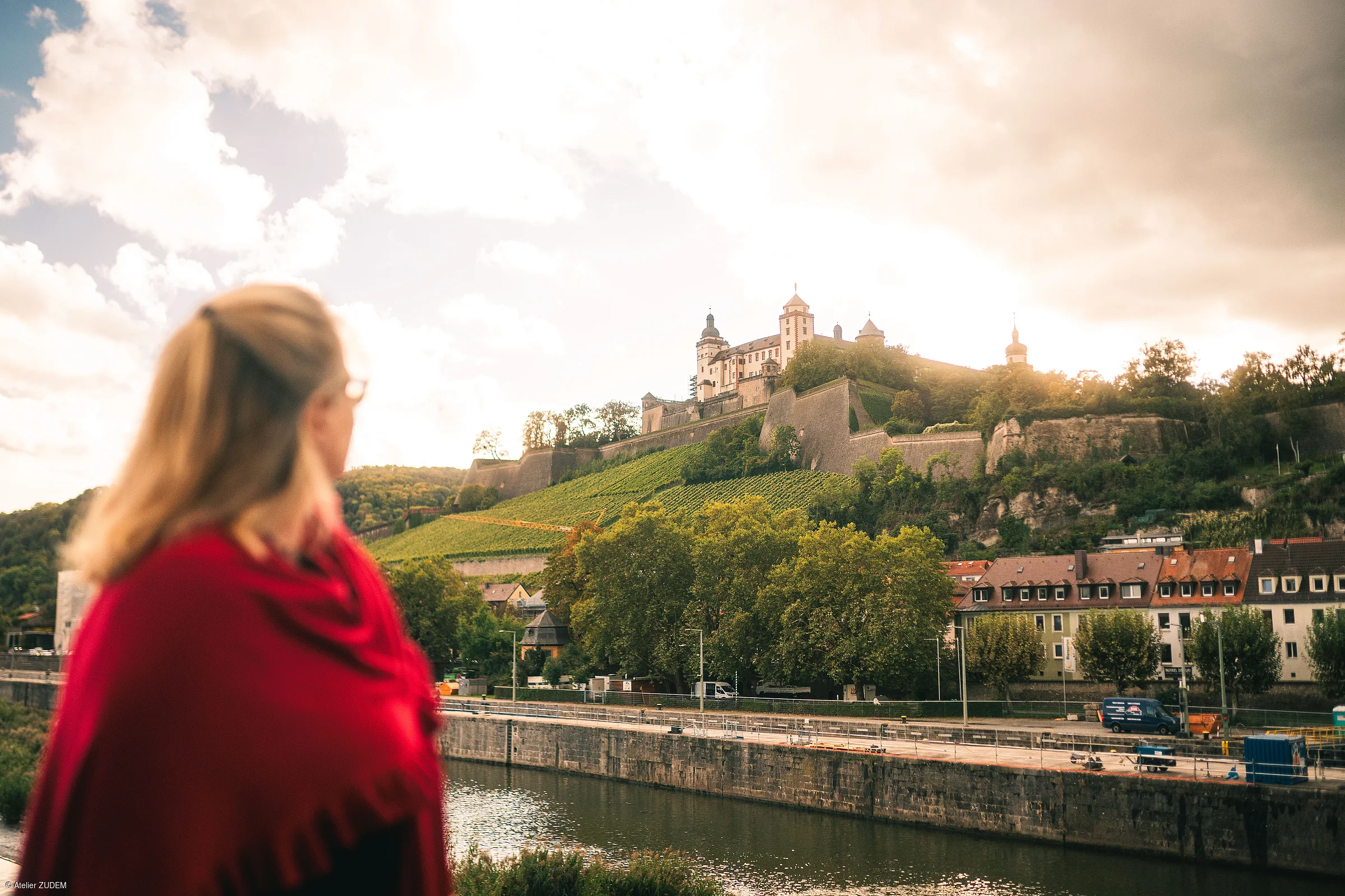 Frau mit rotem Schal blickt auf eine Burg auf einem bewaldeten Hügel über einem Fluss und Häusern.