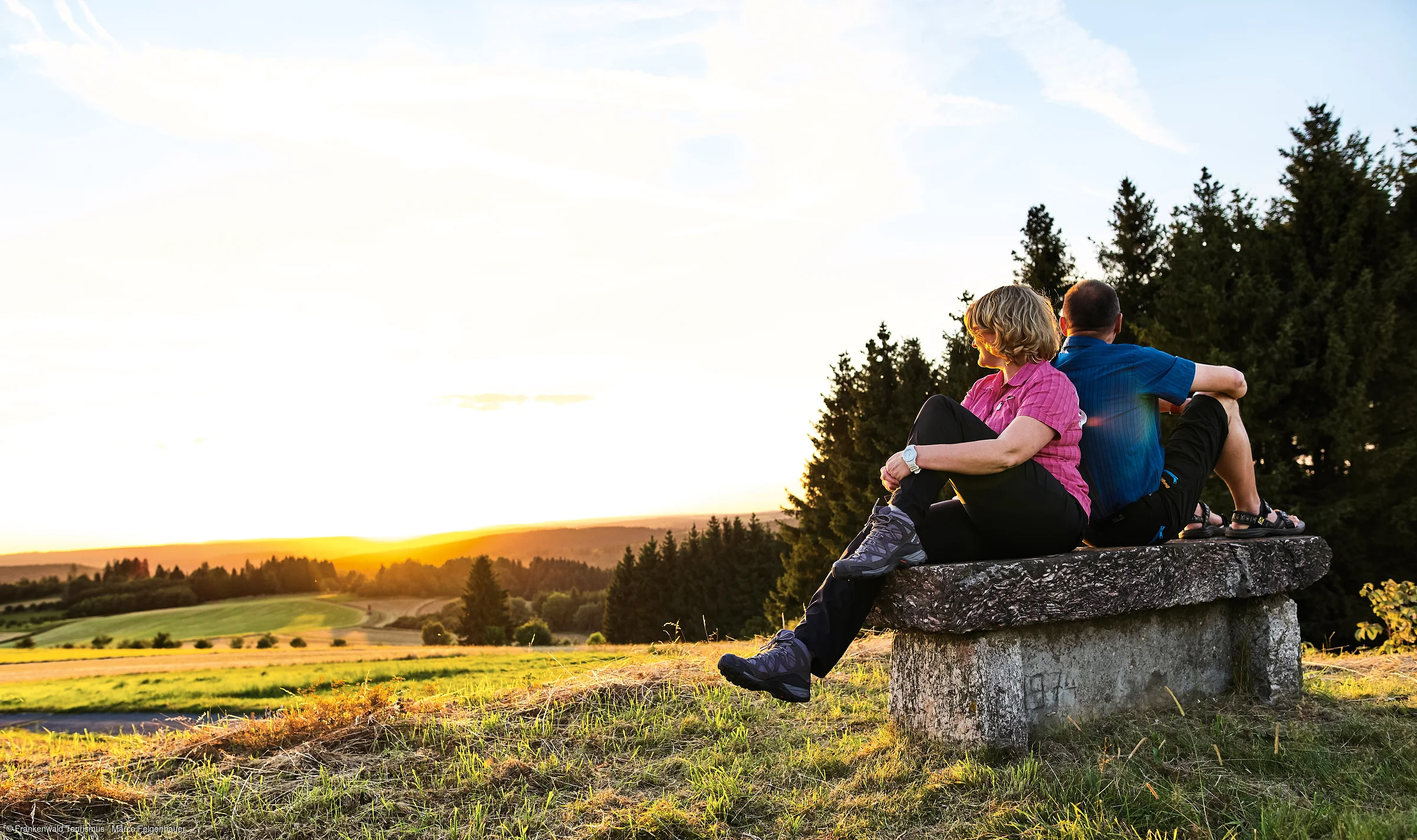 Zwei Personen sitzen auf einer Steinbank und schauen in die untergehende Sonne über einer Landschaft mit Wiesen und Bäumen.