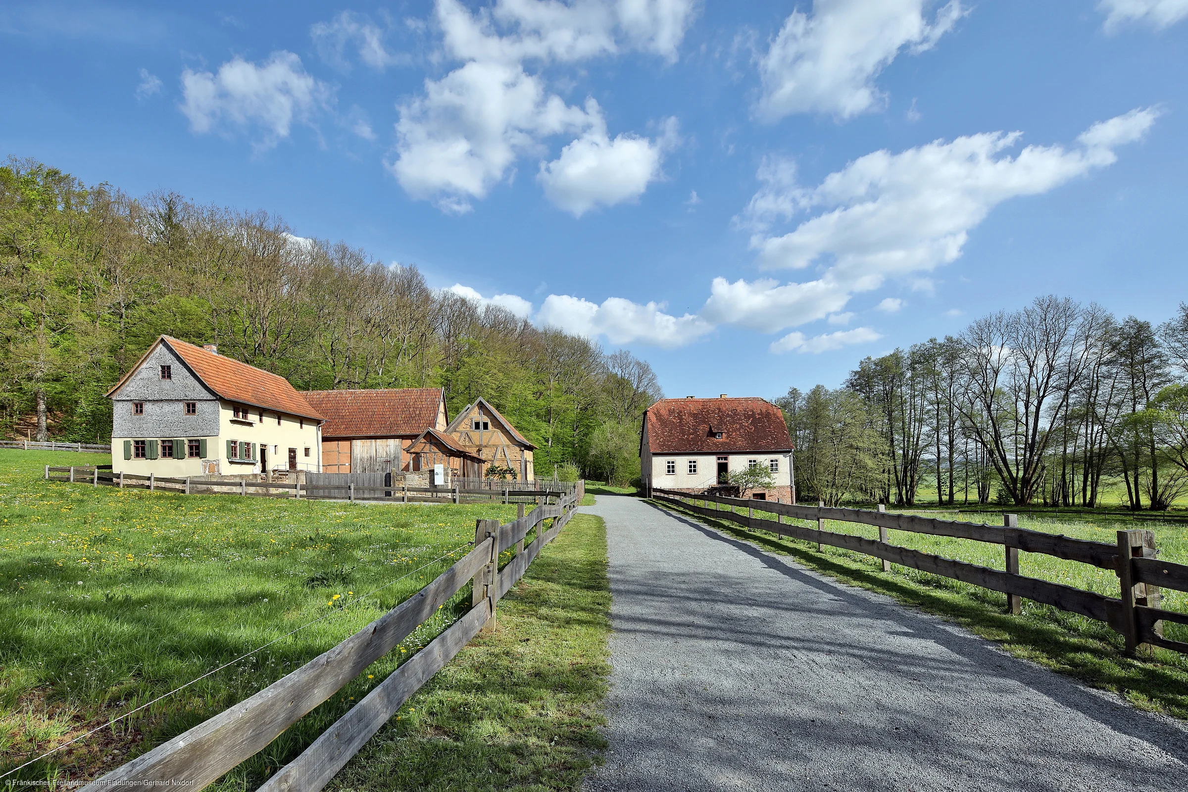 Landstraße mit Holzzäunen führt zu mehreren Fachwerkhäusern vor bewaldetem Hügel unter blauem Himmel mit Wolken