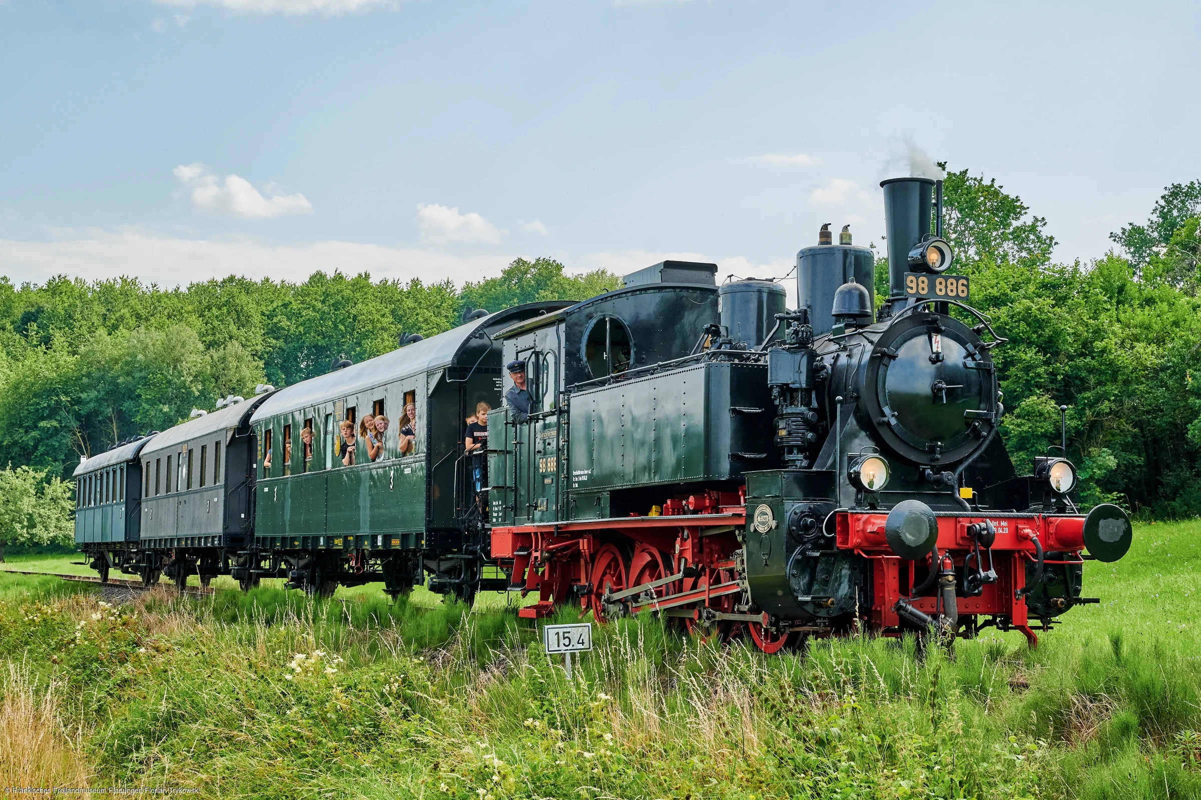 Dampflokomotive mit mehreren Waggons auf Gleisen in grüner Landschaft unter blauem Himmel