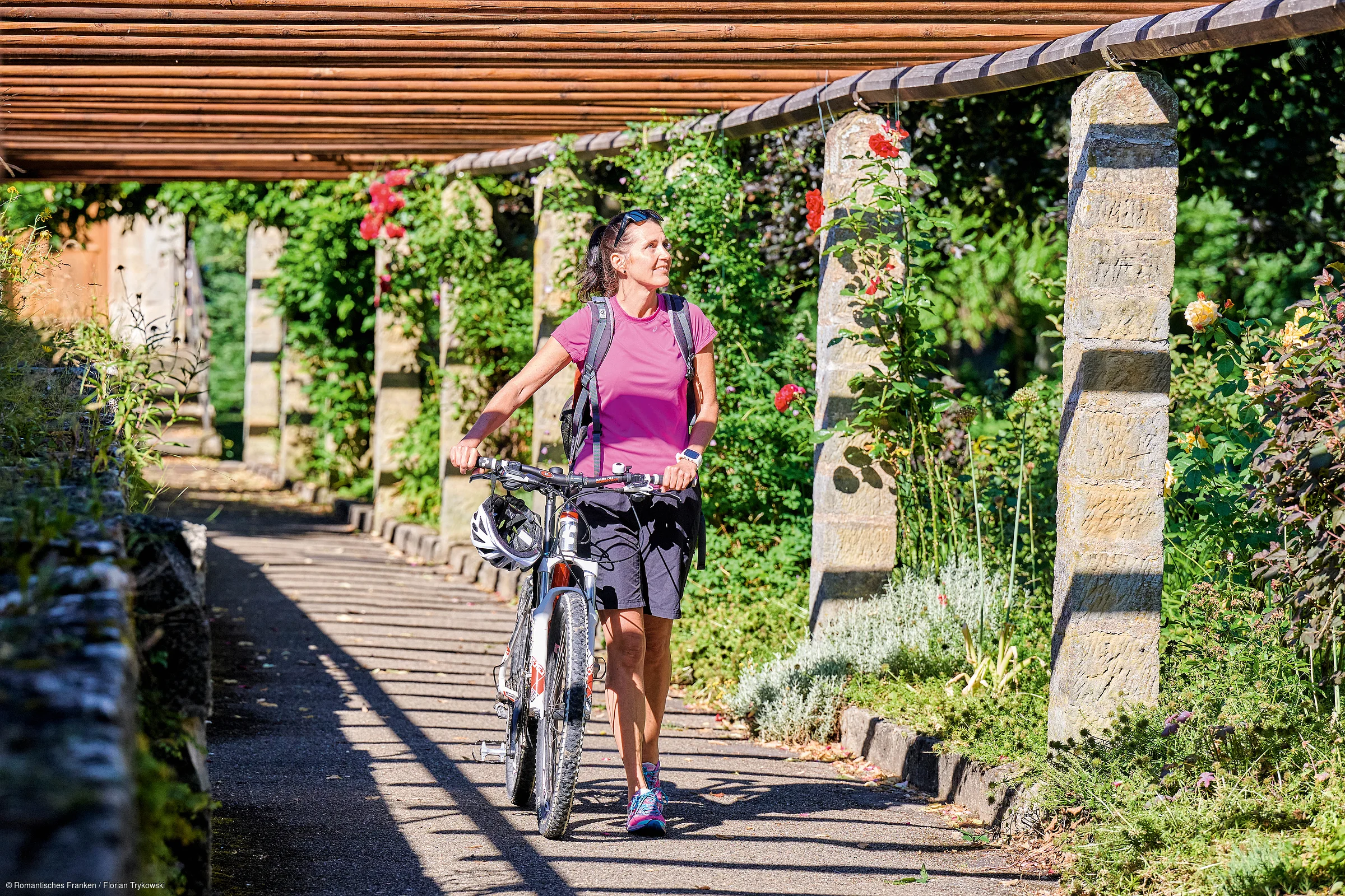 Frau in Sportkleidung schiebt Fahrrad unter Pergola mit Rosen und Säulen entlang eines Weges.