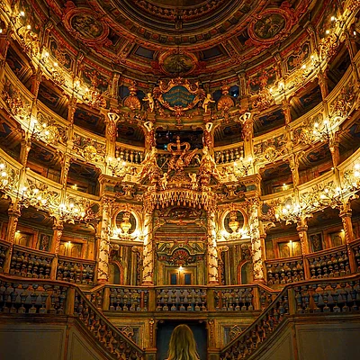 Frau von hinten sitzt in blauem Theatersaal mit reich verziertem, goldbeleuchtetem Zuschauerbereich und Balkon.