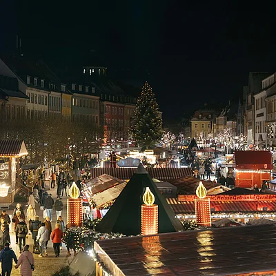 Weihnachtsmarkt bei Nacht mit beleuchteten Ständen, Menschen und einem großen Weihnachtsbaum in der Mitte.