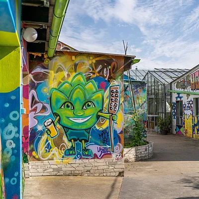 Graffiti mit grünem Comic-Charakter an Hauswand, daneben bunte Wandmalereien und Gewächshaus unter blauem Himmel.