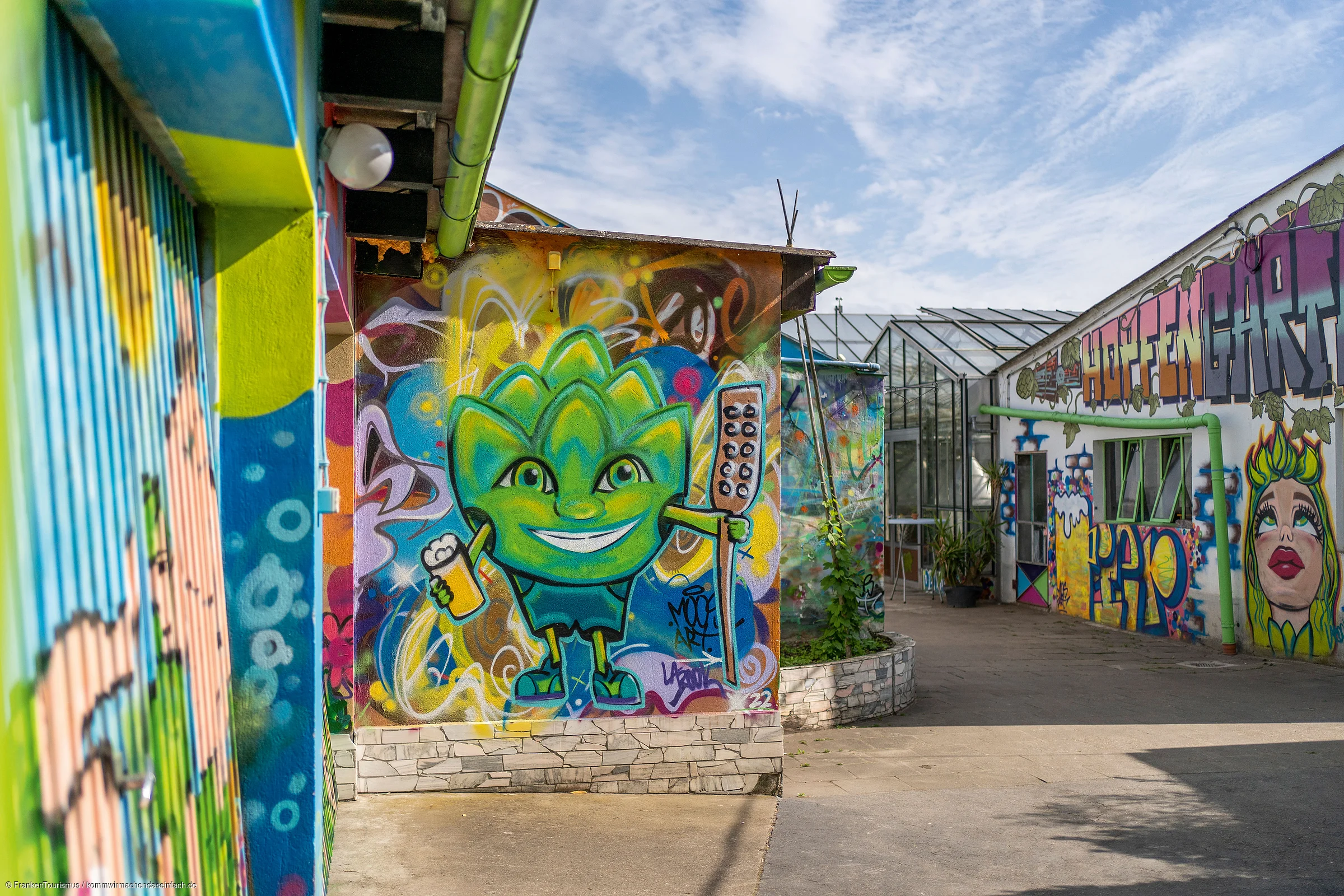 Graffiti mit grünem Comic-Charakter an Hauswand, daneben bunte Wandmalereien und Gewächshaus unter blauem Himmel.