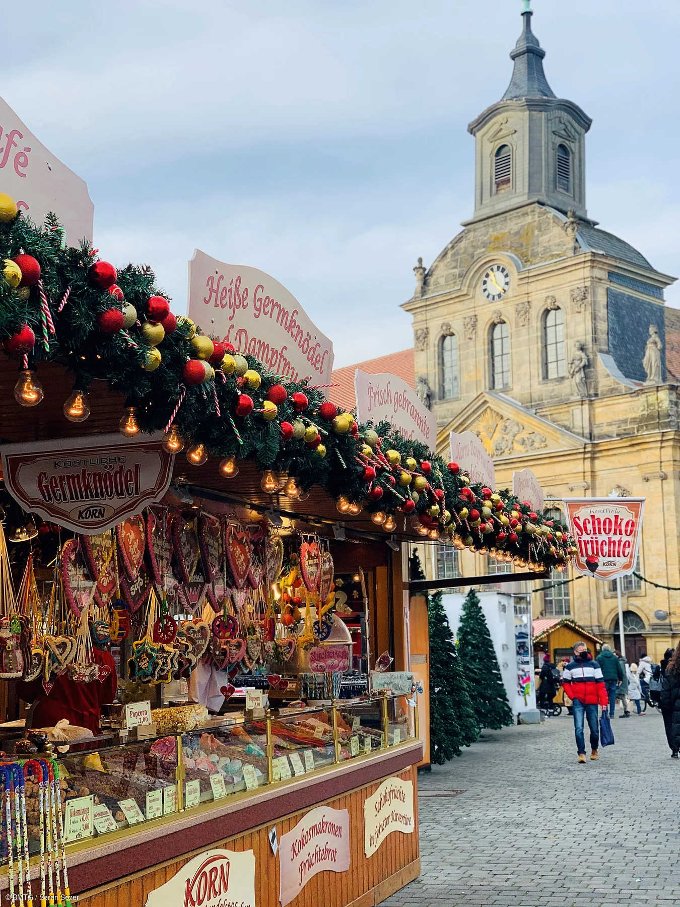 Weihnachtsmarktstand mit Lebkuchenherzen und Dekoration vor historischer Kirche und Passanten auf Pflasterstraße