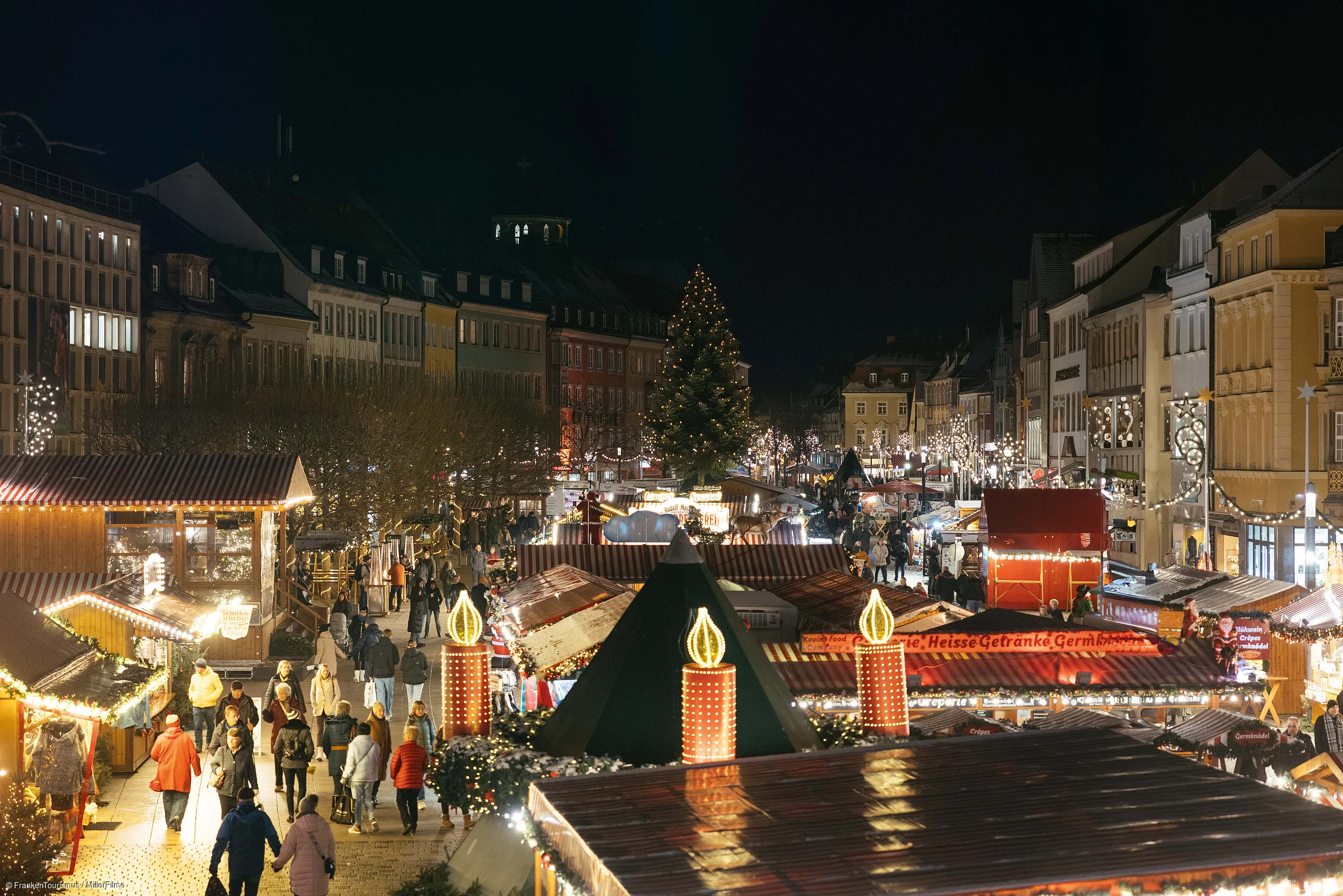Weihnachtsmarkt bei Nacht mit beleuchteten Ständen, Menschen und einem großen Weihnachtsbaum in der Mitte.