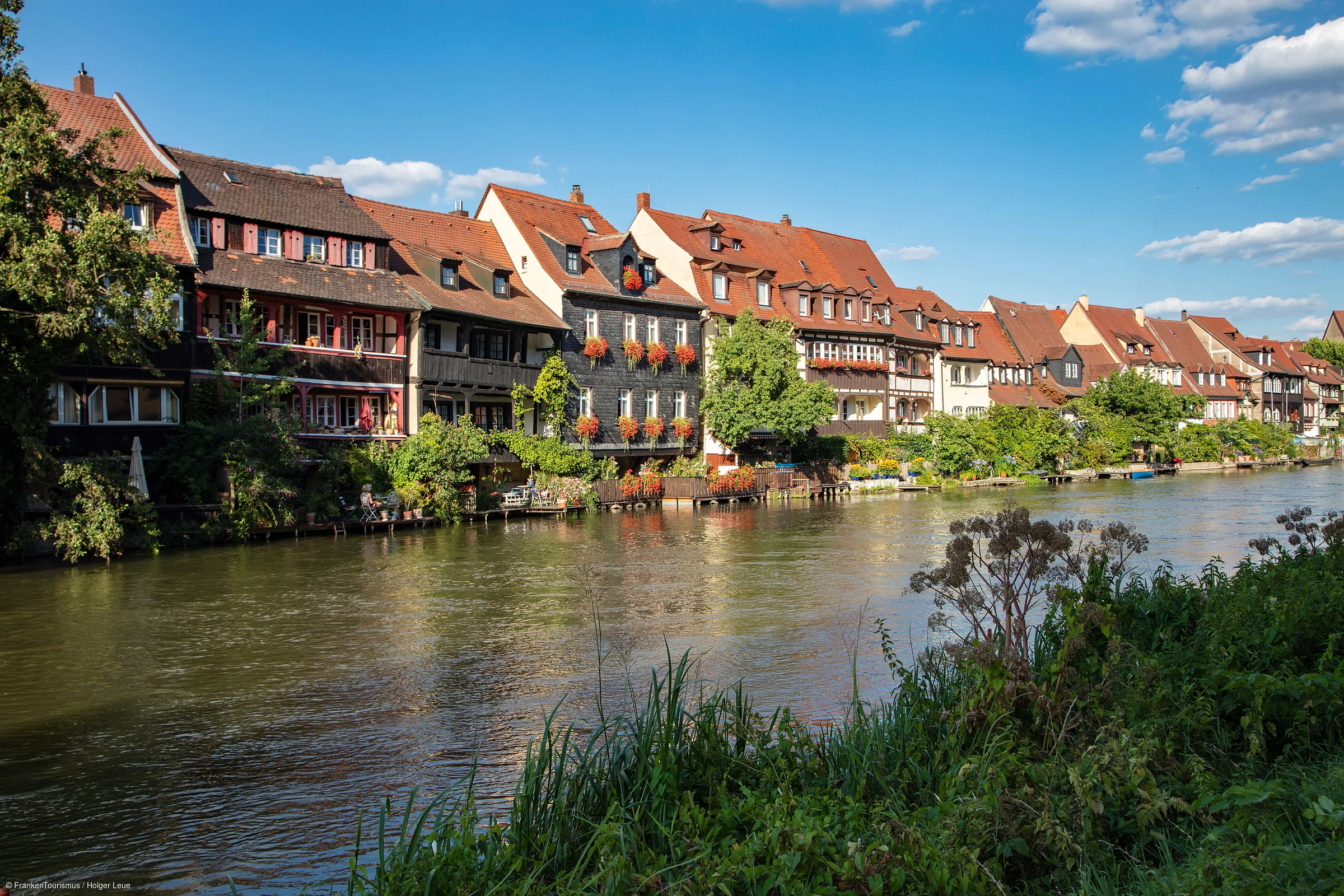 Flussufer mit grüner Vegetation und Fachwerkhäusern mit roten Dächern unter blauem Himmel am Wasser.