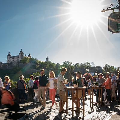 Menschen stehen und gehen auf gepflasterter Brücke bei sonnigem Himmel vor Schloss auf Hügel, Schild Alte Mainmühle sichtbar