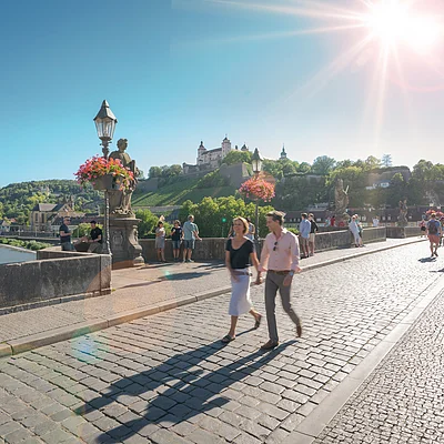 Paar geht auf gepflasterter Brücke mit Blumenampeln, Burg und Fluss im Hintergrund bei Sonnenschein.