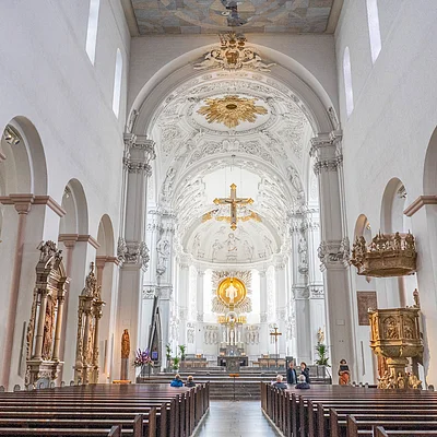 Innenraum einer Kirche mit weißen Wänden, verzierten Altären, Holzbänken und einem goldenen Kreuz über dem Altar.