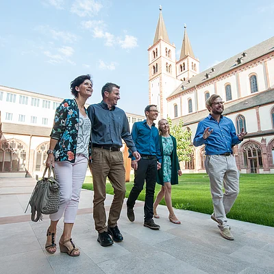 Fünf Personen gehen lachend auf einem gepflasterten Weg neben einer historischen Kirche mit zwei Türmen.