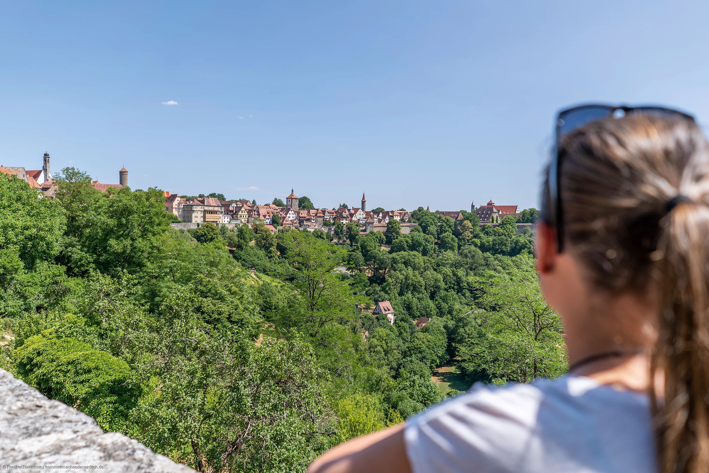 Frau mit Pferdeschwanz und Sonnenbrille blickt auf bewaldeten Hügel und Stadt mit Fachwerkhäusern bei klarem Himmel.
