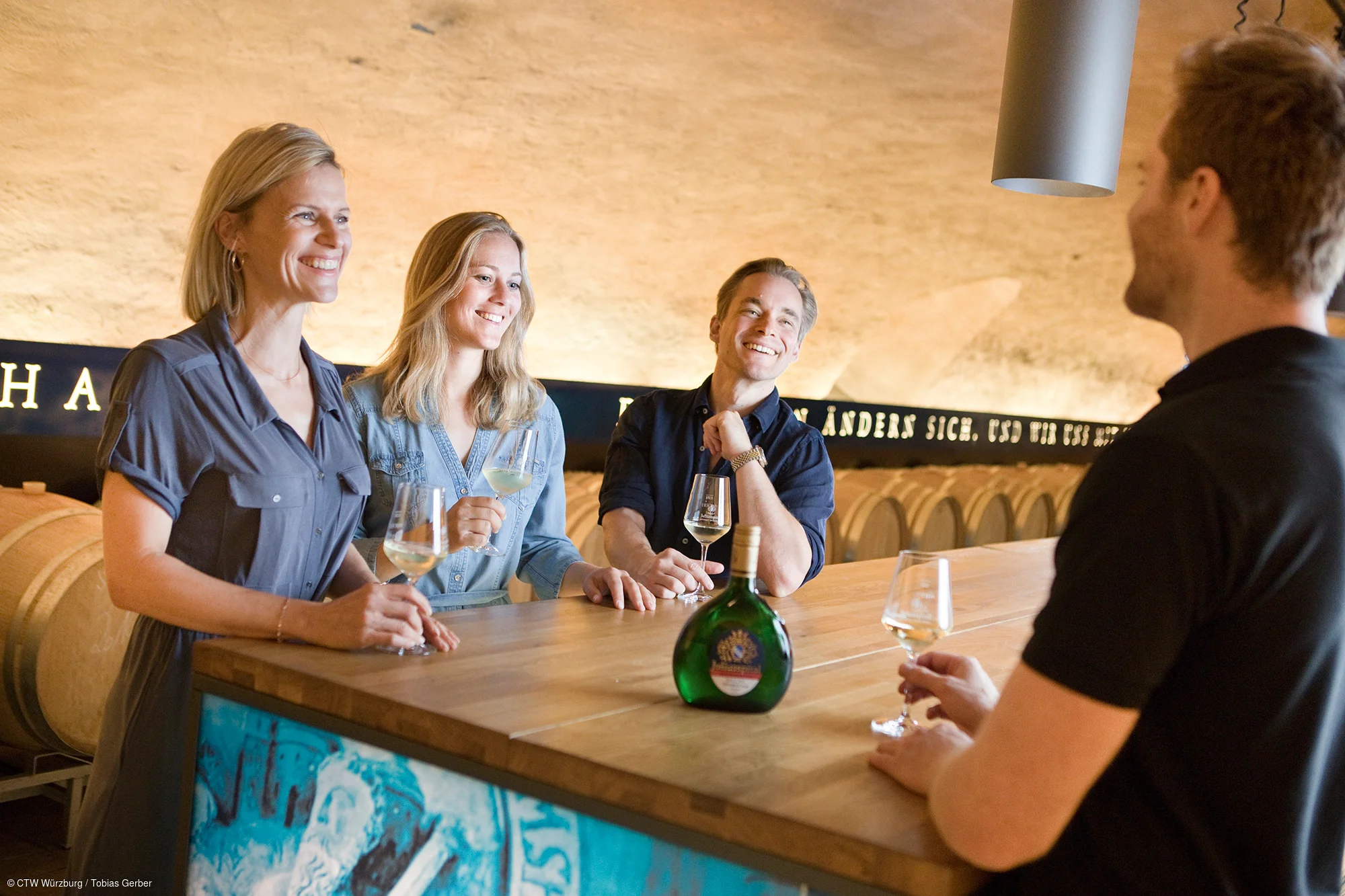 Drei Personen stehen an einem Holztresen mit Weingläsern, eine vierte Person schenkt ihnen Wein in einem Keller mit Weinfässern.