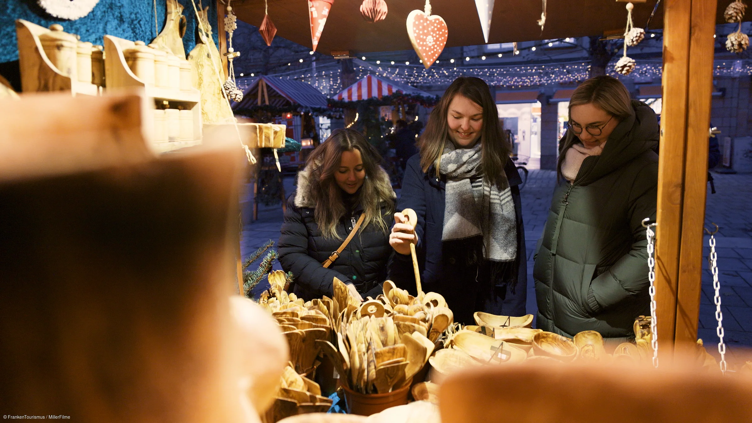 Drei Frauen in Winterjacken betrachten Holzlöffel und -schalen an einem Marktstand mit Weihnachtsdekoration.