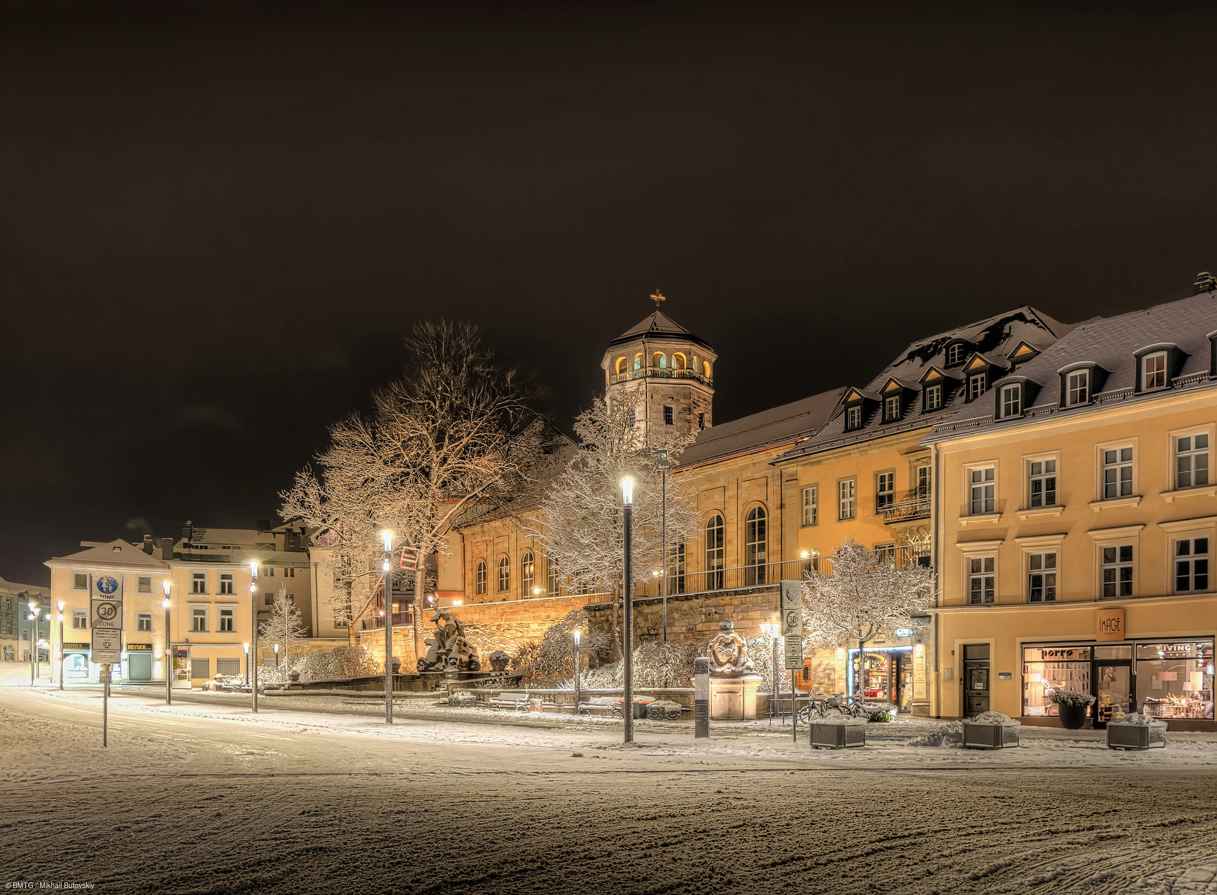 Schneebedeckte Straße mit beleuchteten Gebäuden und Bäumen bei Nacht in einer Stadt.
