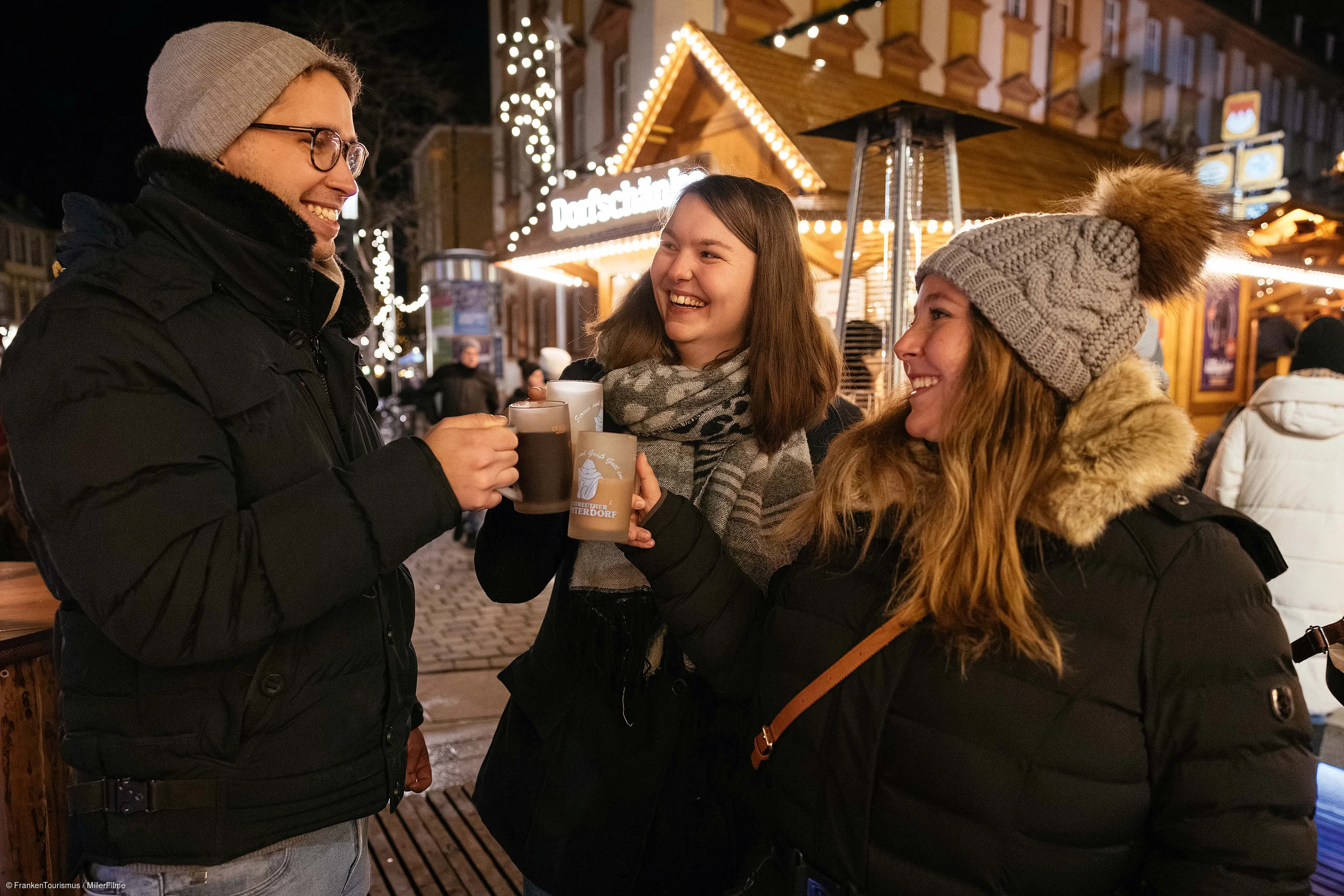 Drei Personen stoßen mit Tassen bei einem beleuchteten Weihnachtsmarktstand im Freien an.