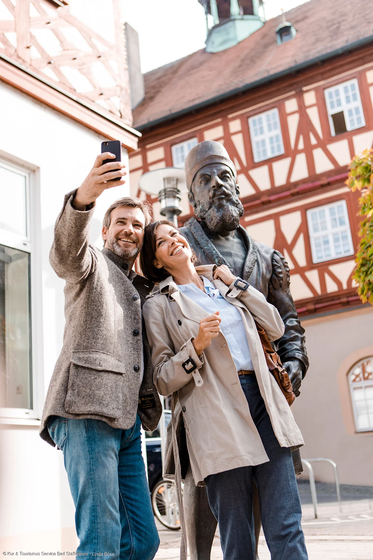 Paar macht Selfie vor einer historischen Statue in einer Altstadt mit Fachwerkhäusern.
