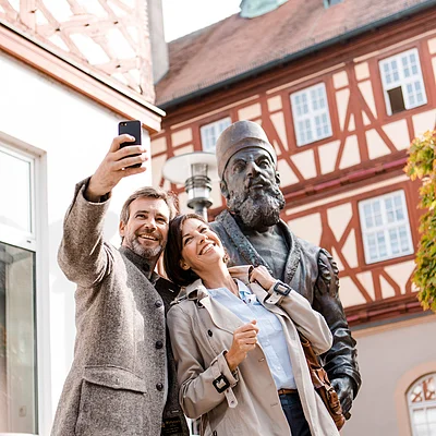 Paar macht Selfie vor einer historischen Statue in einer Altstadt mit Fachwerkhäusern.