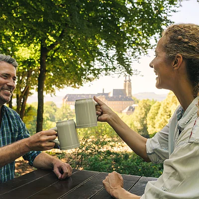 Zwei Personen stoßen mit Bierkrügen an einem Holztisch im Freien an, im Hintergrund Bäume und ein Gebäude.