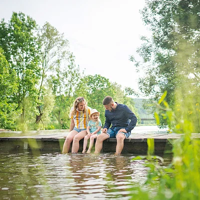 Eine Familie sitzt auf einem Steg am Wasser, umgeben von Bäumen und Pflanzen.
