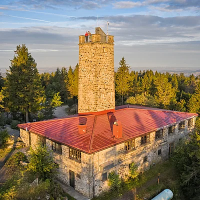 Steinerner Aussichtsturm mit rotem Dach umgeben von Nadelbäumen bei bewölktem Himmel.