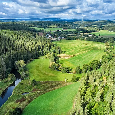 Landschaft mit Wäldern, Wiesen, Fluss, Windrädern und einzelnen Häusern unter bewölktem Himmel.