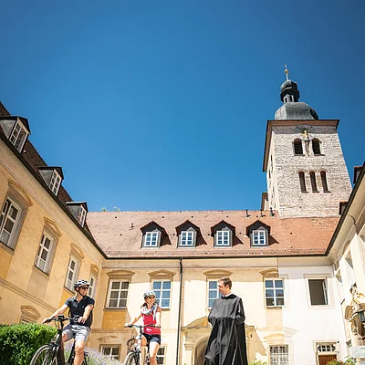 Zwei Radfahrer und eine Person in schwarzer Robe vor einem historischen Gebäude mit Turm.
