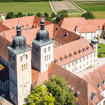 Luftaufnahme eines Klosters mit roten Dächern, zwei Türmen und umliegenden Gebäuden. Im Hintergrund sind Felder zu sehen.