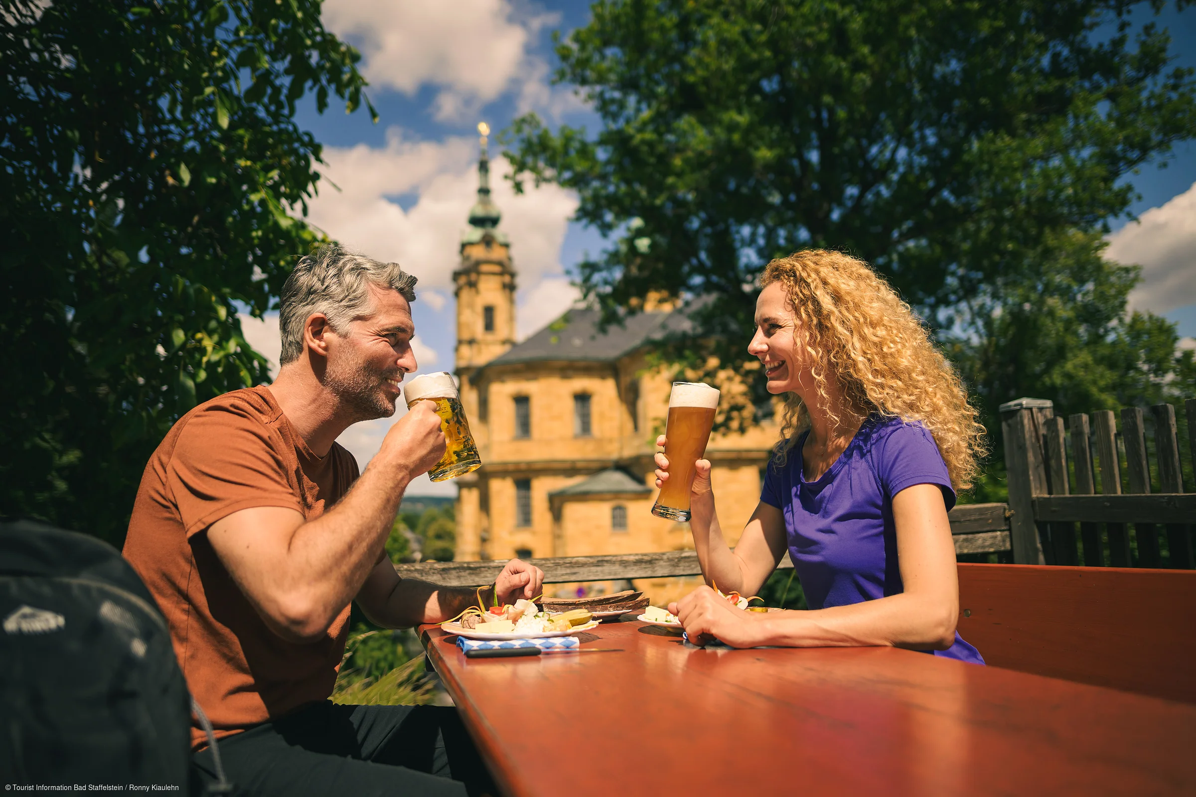Mann und Frau sitzen draußen an Holztisch und trinken Bier vor historischer Kirche bei Sonnenschein.