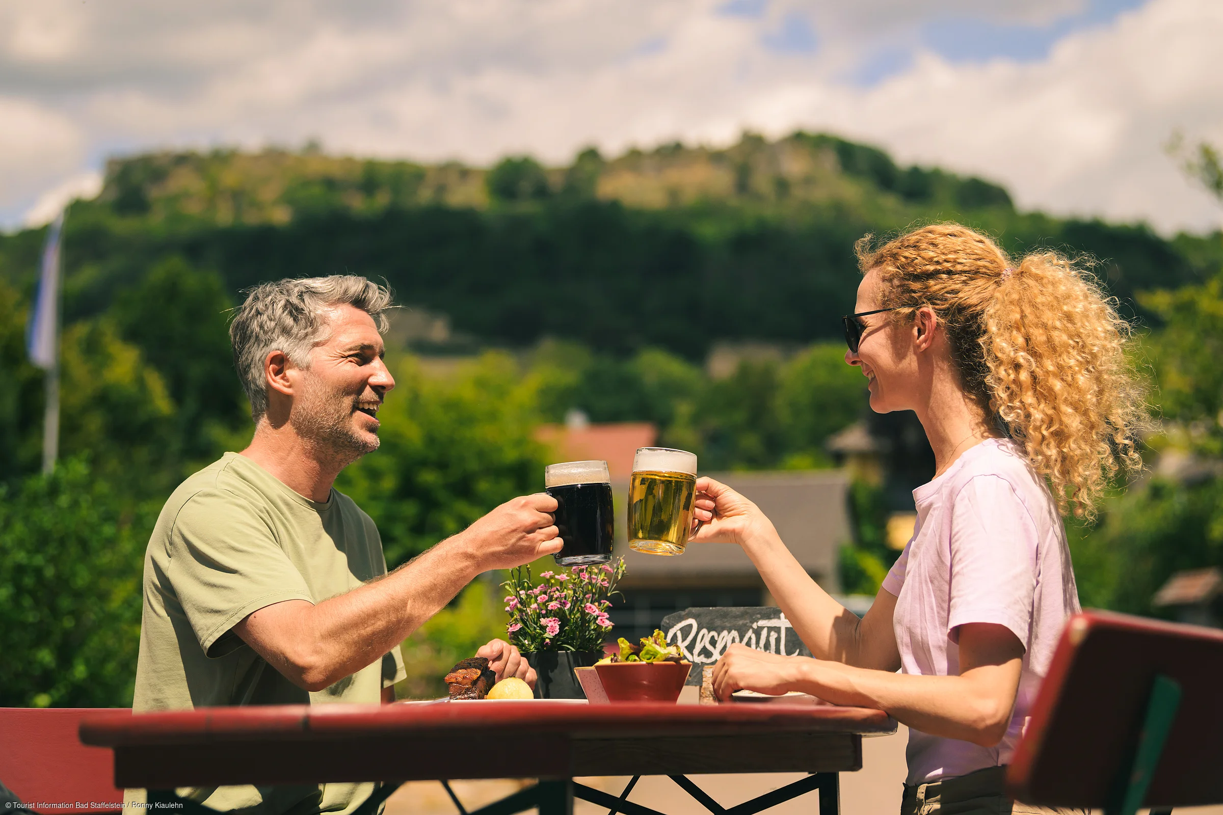Mann und Frau stoßen mit Biergläsern an, sitzen draußen an einem Tisch mit Pflanzen und Berg im Hintergrund.