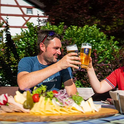 Mann und Frau stoßen mit Biergläsern an, davor Brett mit Käse, Wurst und Brot auf einem Tisch.