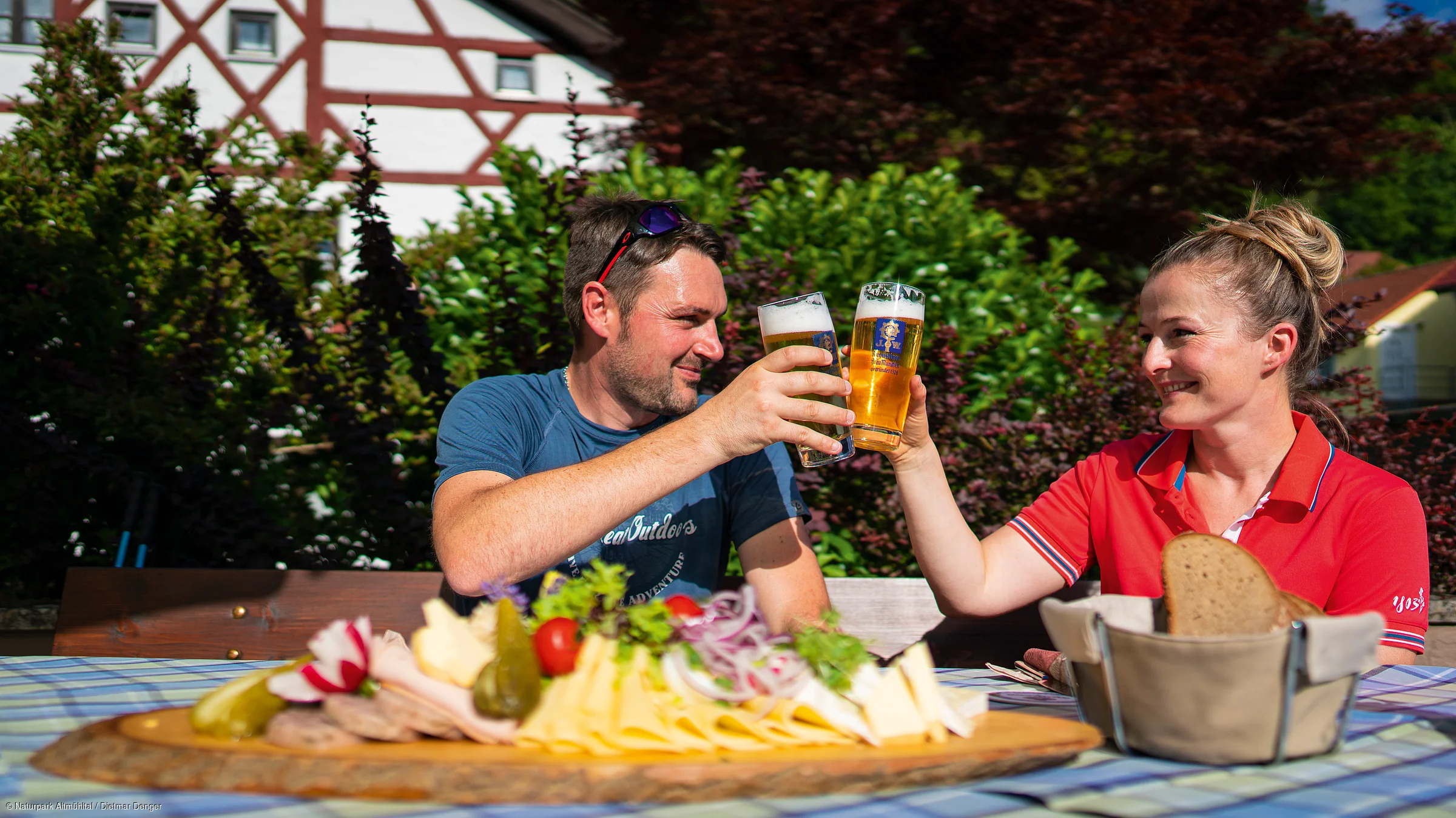Mann und Frau stoßen mit Biergläsern an, davor Brett mit Käse, Wurst und Brot auf einem Tisch.