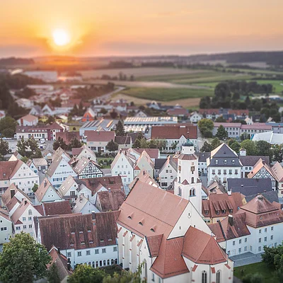 Luftaufnahme einer Stadt mit roten Dächern bei Sonnenuntergang und Feldern im Hintergrund.