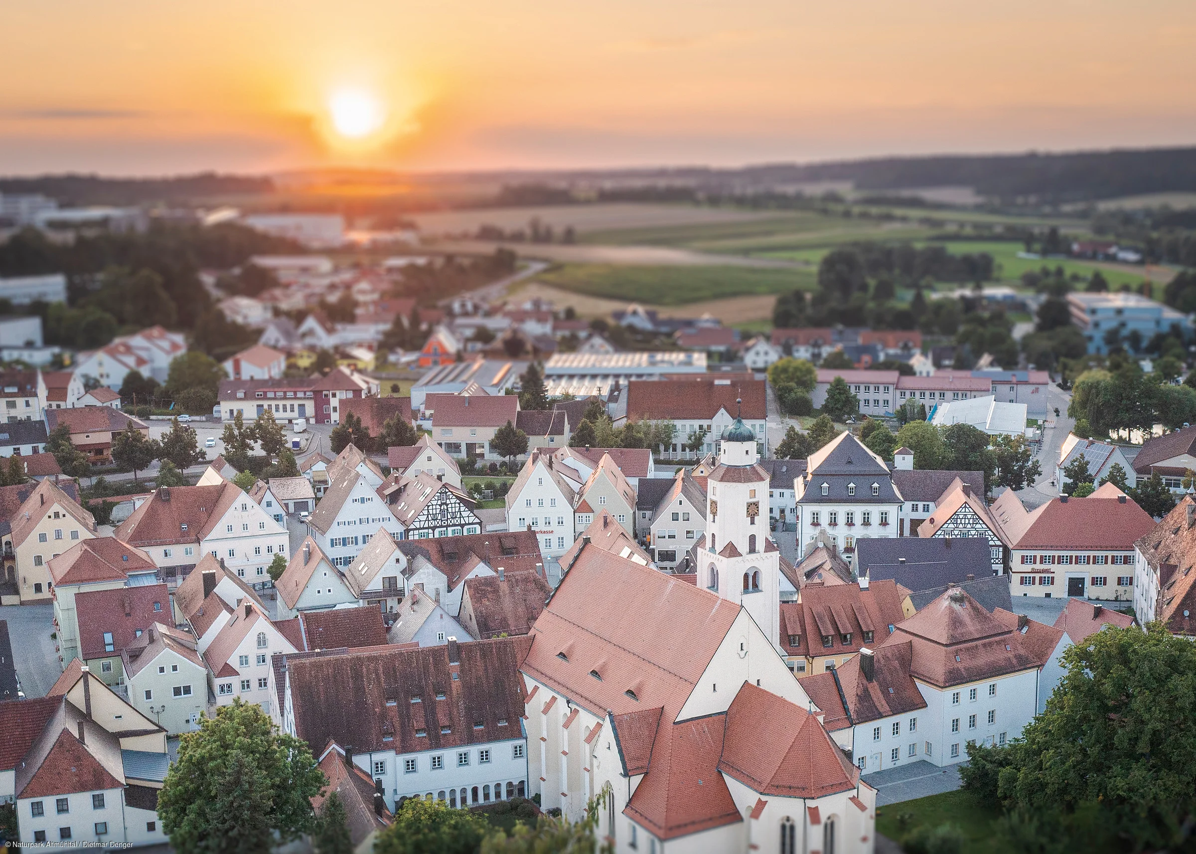 Luftaufnahme einer Stadt mit roten Dächern bei Sonnenuntergang und Feldern im Hintergrund.