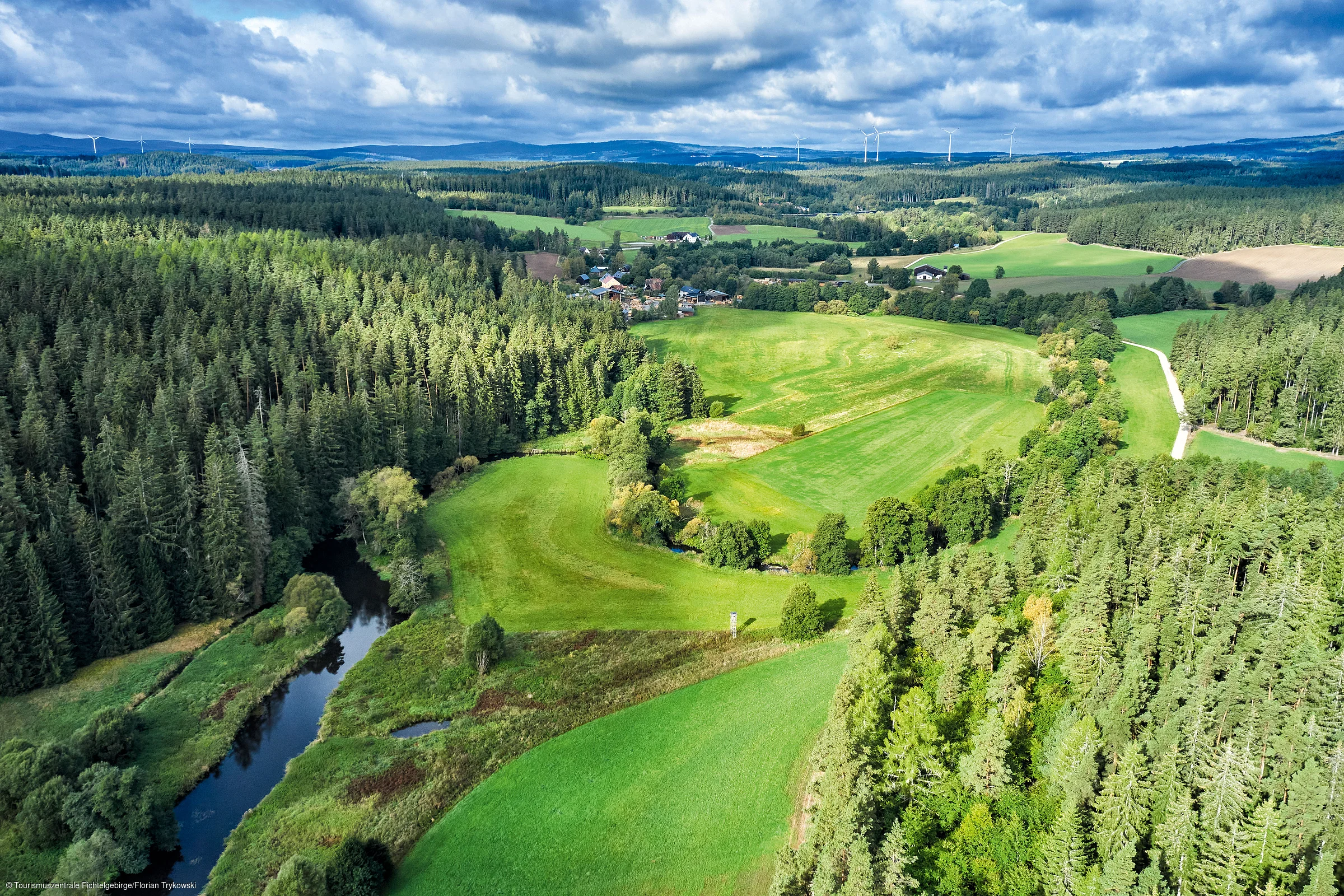Landschaft mit Wäldern, Wiesen, Fluss, Windrädern und einzelnen Häusern unter bewölktem Himmel.