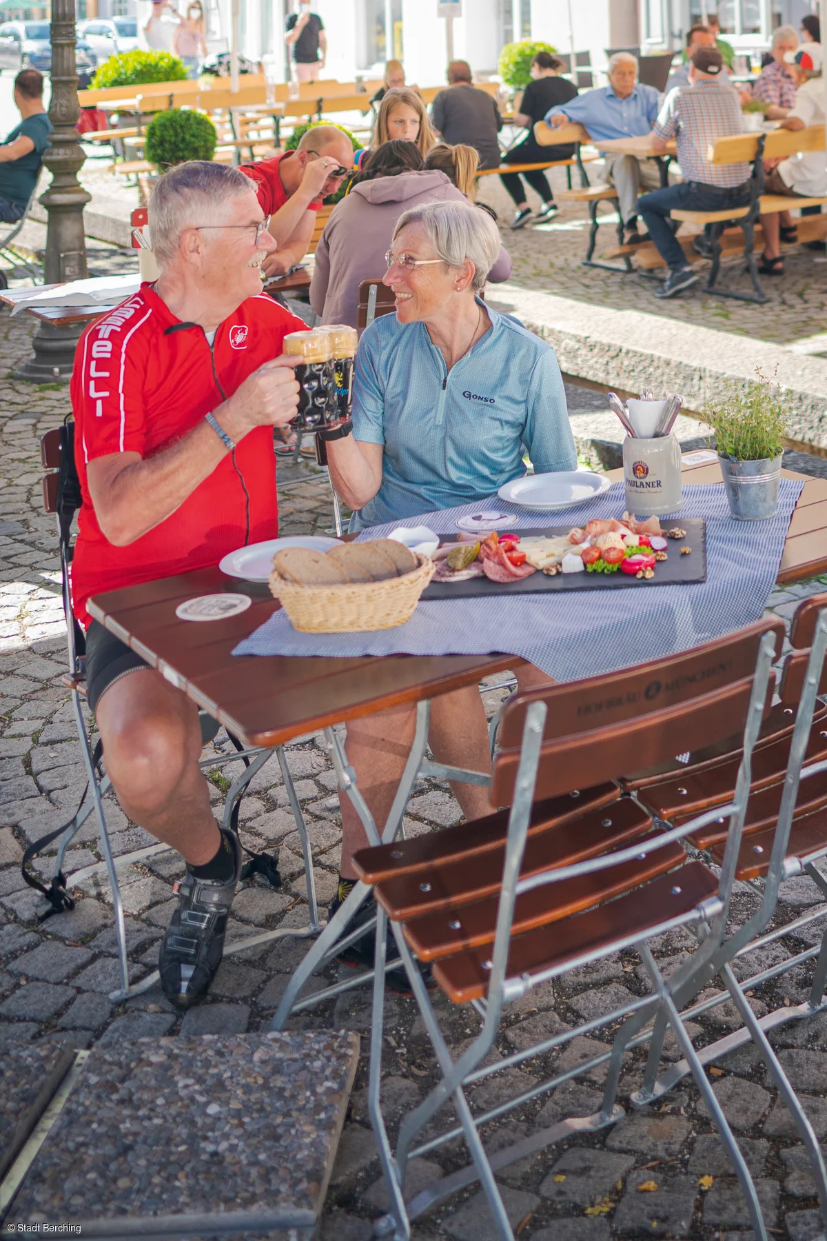 Ein älteres Paar sitzt an einem Tisch im Freien, stößt mit Bierkrügen an und hat eine Brotzeitplatte vor sich.