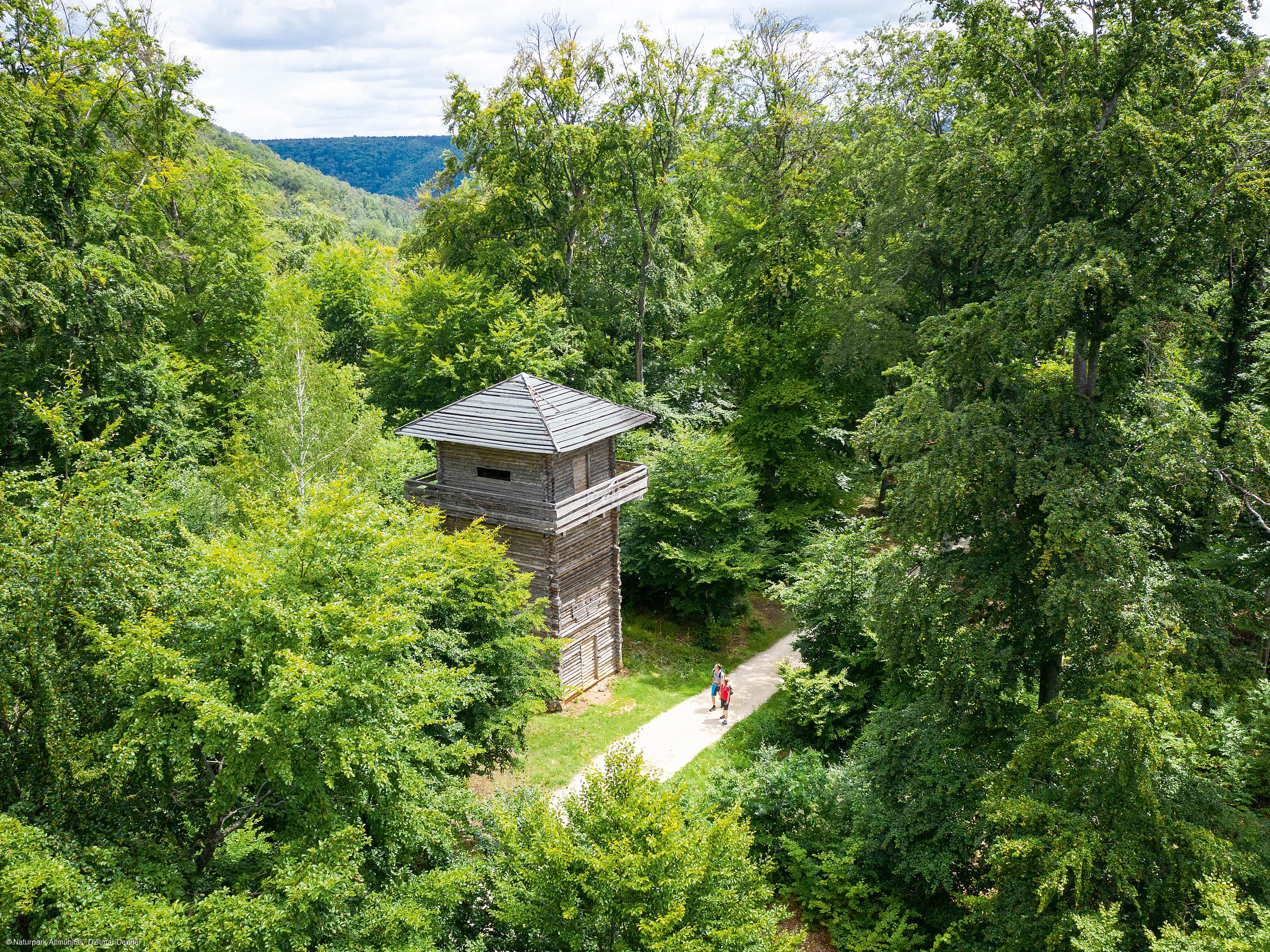 Holzturm neben Wanderweg im dichten grünen Wald mit zwei Personen auf dem Weg sichtbar