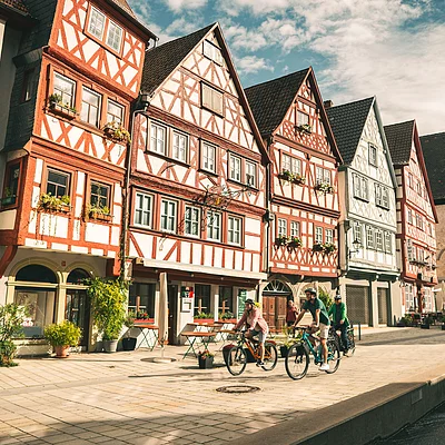Fachwerkhäuser an gepflasterter Straße mit Radfahrern und Wasserkanal bei bewölktem Himmel.