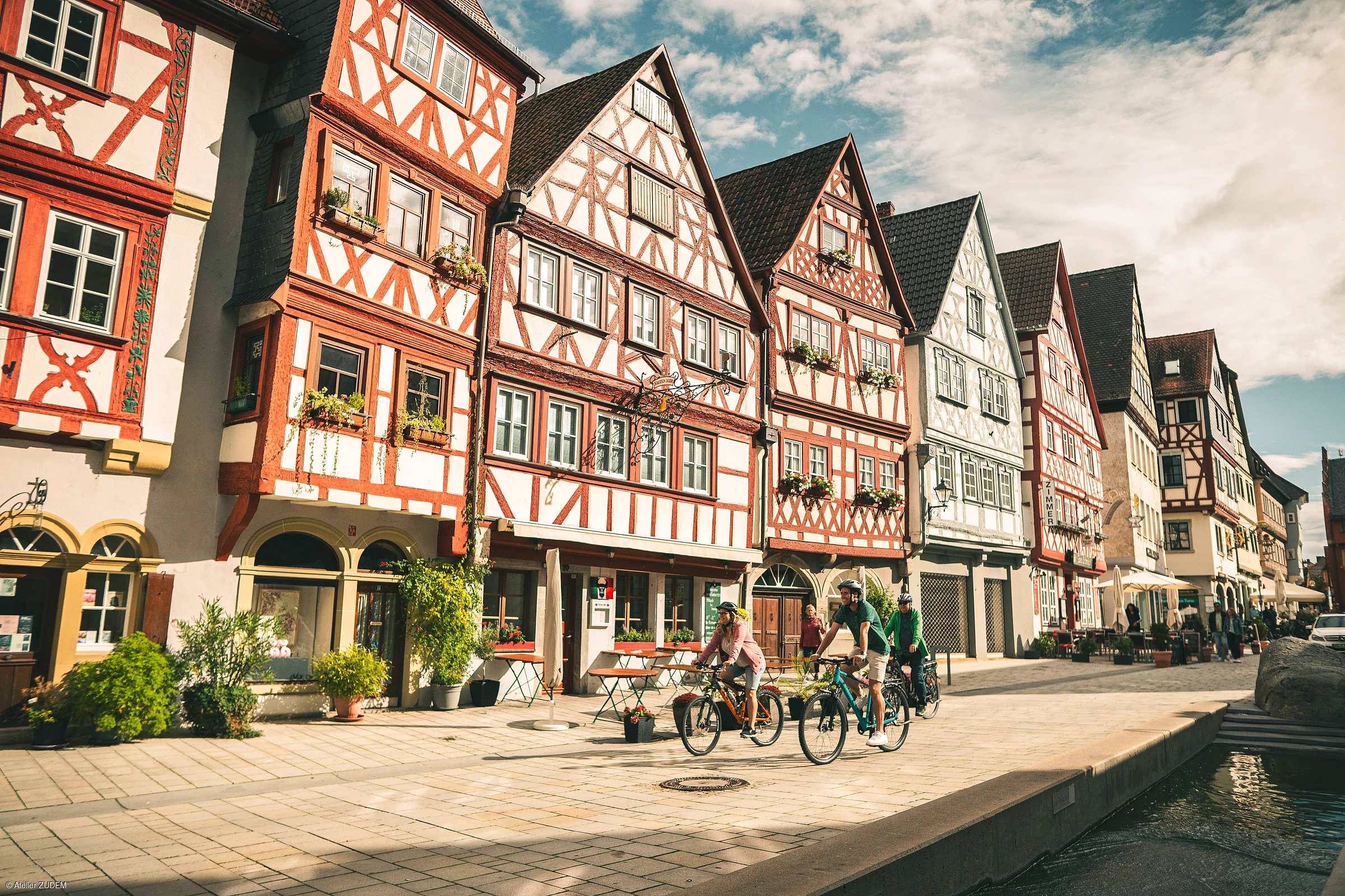 Fachwerkhäuser an gepflasterter Straße mit Radfahrern und Wasserkanal bei bewölktem Himmel.