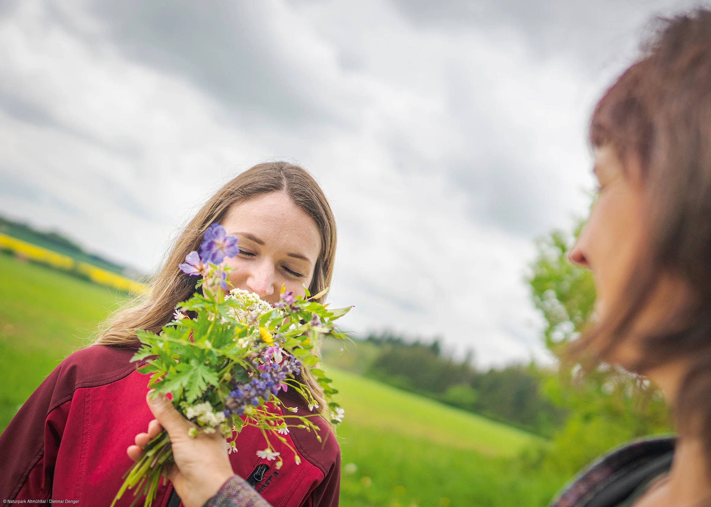 Frau riecht an einem Strauß mit Kräutern, der ihr von einer anderen Person im Freien überreicht wird.