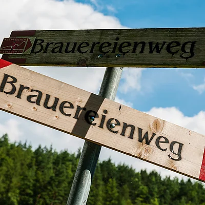 Zwei hölzerne Wegweiser mit der Aufschrift „Brauereienweg“ vor blauem Himmel und Wald im Hintergrund.